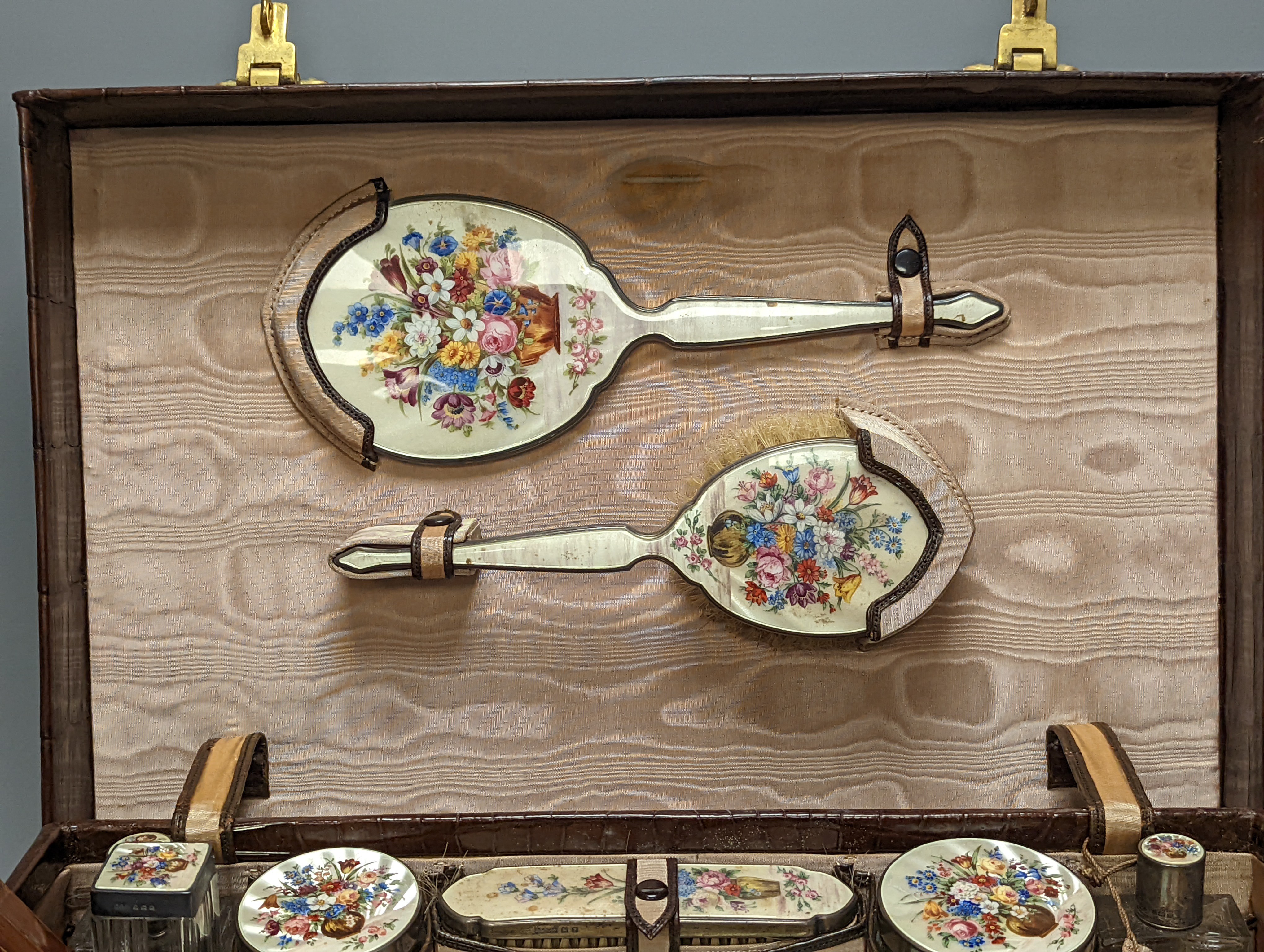 A 1930's crocodile skin travelling toilet case, with ten silver and floral enamel mounted accoutrements, Henry Clifford Davis, Birmingham, 1935,6, together with five other silver and enamel brushes and mirror etc and a c